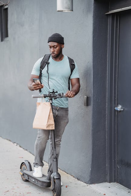A young man with dark skin, wearing a black beanie and light blue T-shirt, is standing outdoors against a gray wall, holding a smartphone with one hand and gripping the handlebar of an electric scoote
