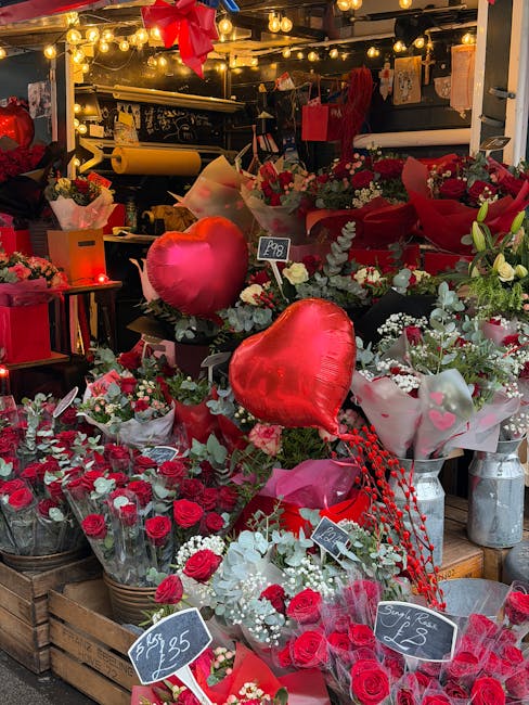 A vibrant floral display at Sidcup Florist, featuring an assortment of red roses, white and green foliage, and heart-shaped foil balloons, arranged in baskets and wrapped in decorative paper. The scen